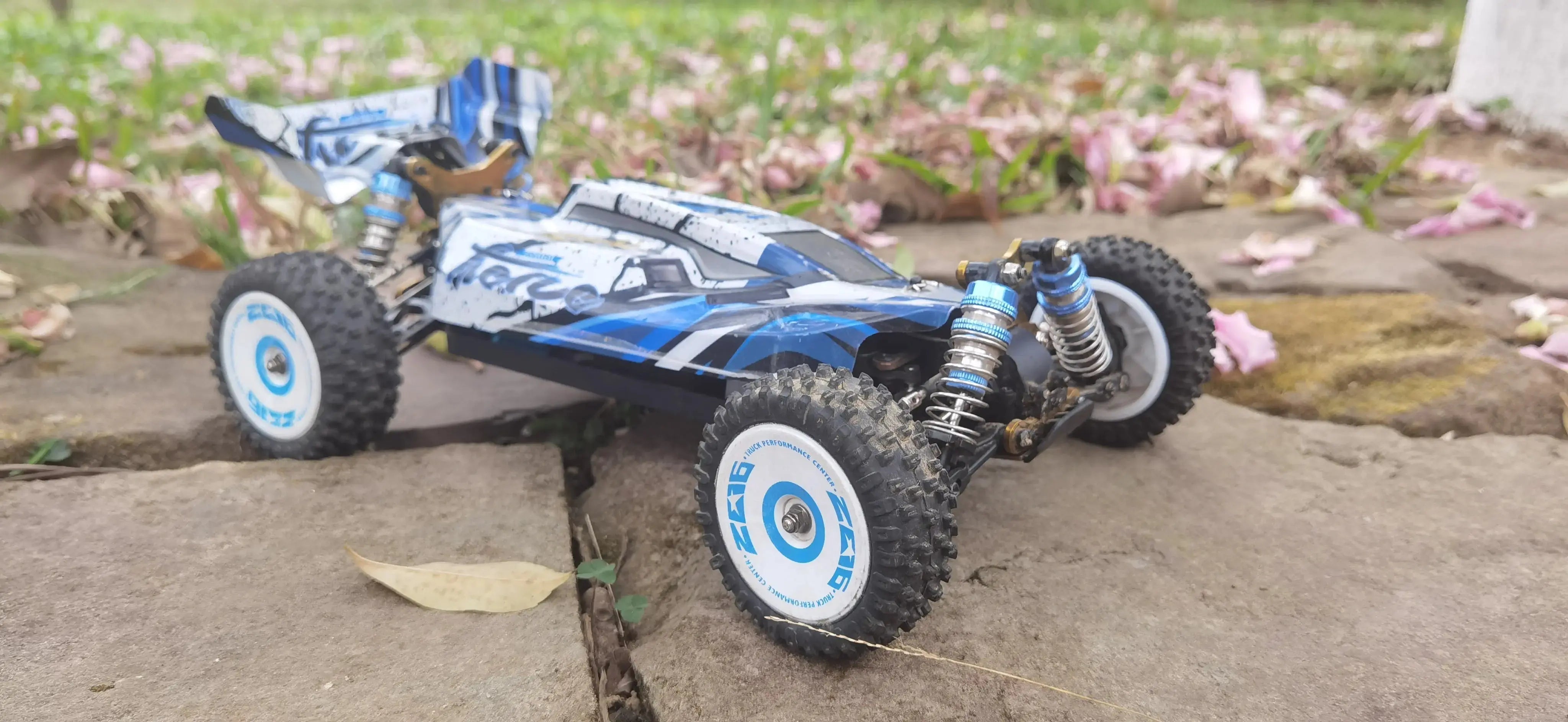 A blue and white 1:10 scale RC Drift Car positioned on a rough gravel surface ideal for drifting, with a springtime outdoor backdrop of scattered petals.