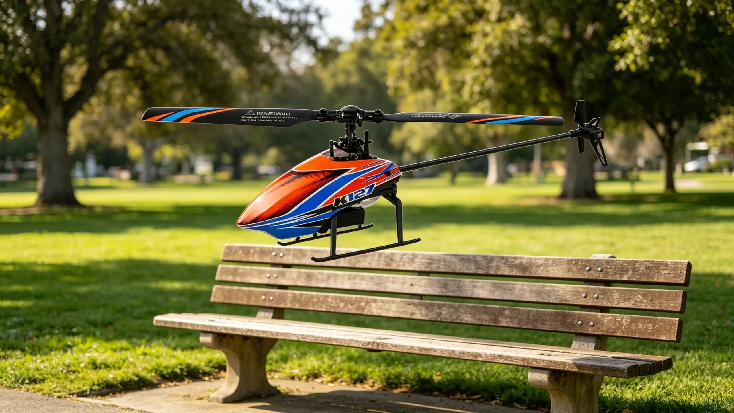 A red, blue, and orange remote control helicopter sits on a grassy field in a park, with a shallow depth of field blurring the background to emphasize the aircraft.