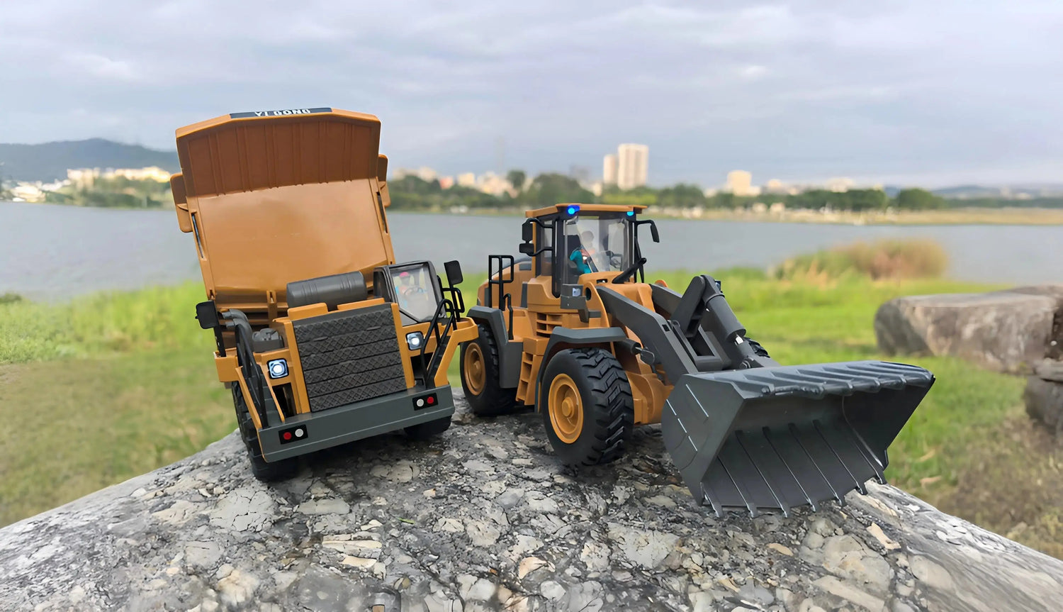 An orange and a black detailed construction vehicle toy models are positioned on outdoor rocks, with a dump truck on the left and a front loader on the right, against a blurred background of a river and city skyline.